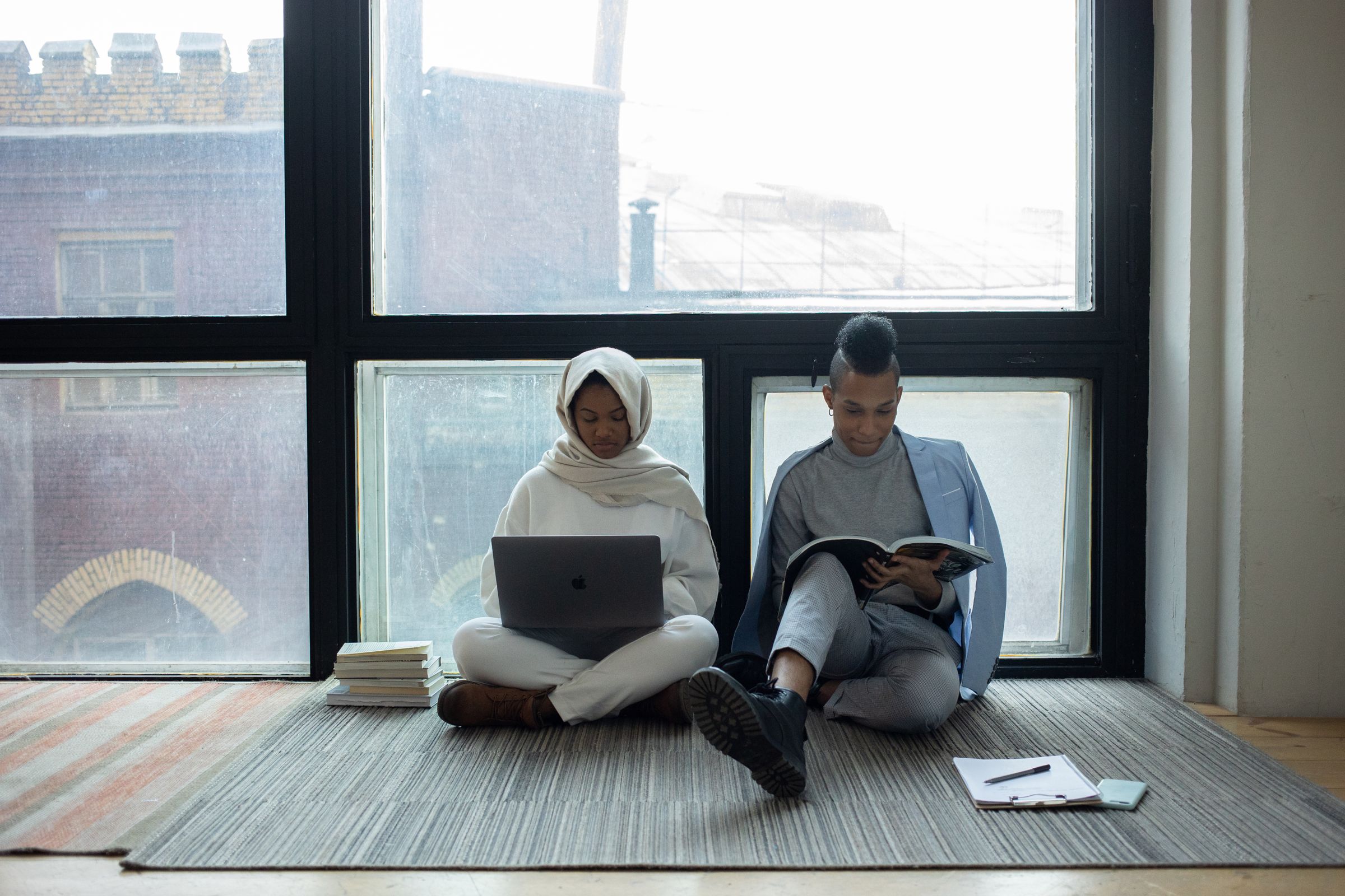 Two students studying beside a tall window, one with a laptop and one with a book.