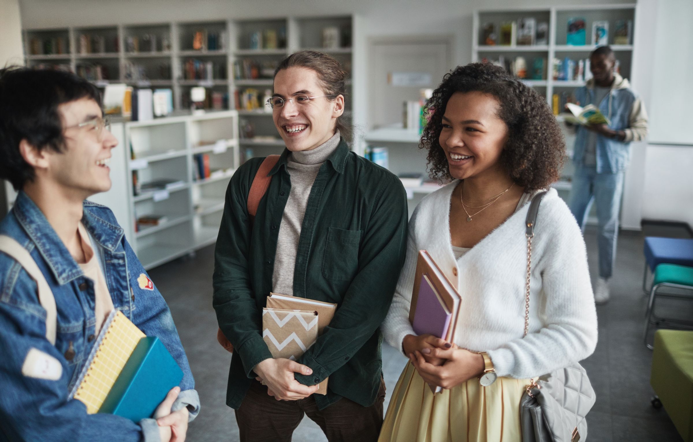 Three students laughing together in a bright school library, holding their books.